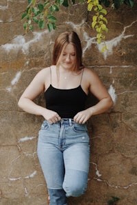 a woman leaning against a wall in jeans and a black tank top