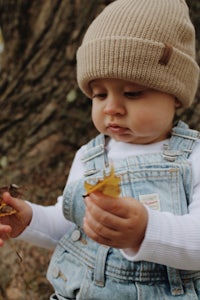 a baby wearing overalls and a beanie holding a leaf