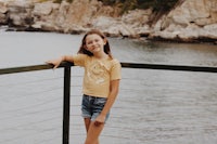 a girl wearing a yellow t - shirt standing on a railing near a body of water