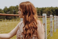 a woman with long red hair standing in a field next to a fence