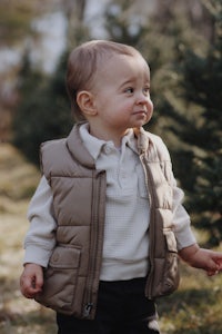 a baby boy in a tan vest standing in front of a christmas tree