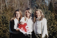 three women pose with a baby in front of a christmas tree farm